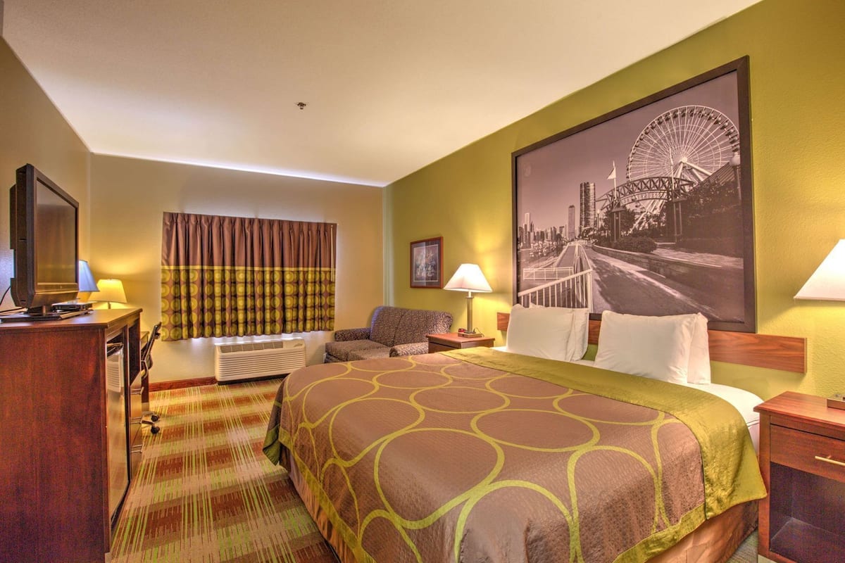 Ein Hotelzimmer mit Bett, Schreibtisch mit Fernseher, Sessel, Fenster mit Vorhängen, Wandlampe und großem Stadtplakat.