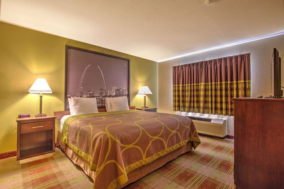 Ein Hotelzimmer mit einem Bett, zwei Nachttischen, einem Fenster mit Vorhängen, einem an der Wand montierten Fernseher und einem Stadtbild-Poster.