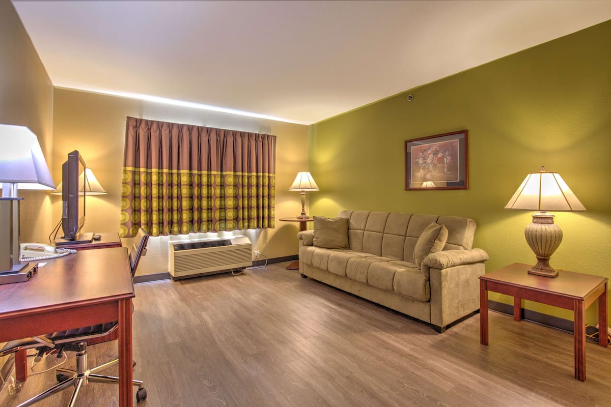 Ein Hotelzimmer mit Holzboden, einem beigefarbenen Sofa, einem Schreibtisch mit Computer und Telefon, zwei Lampen und einem fenster mit Vorhängen.