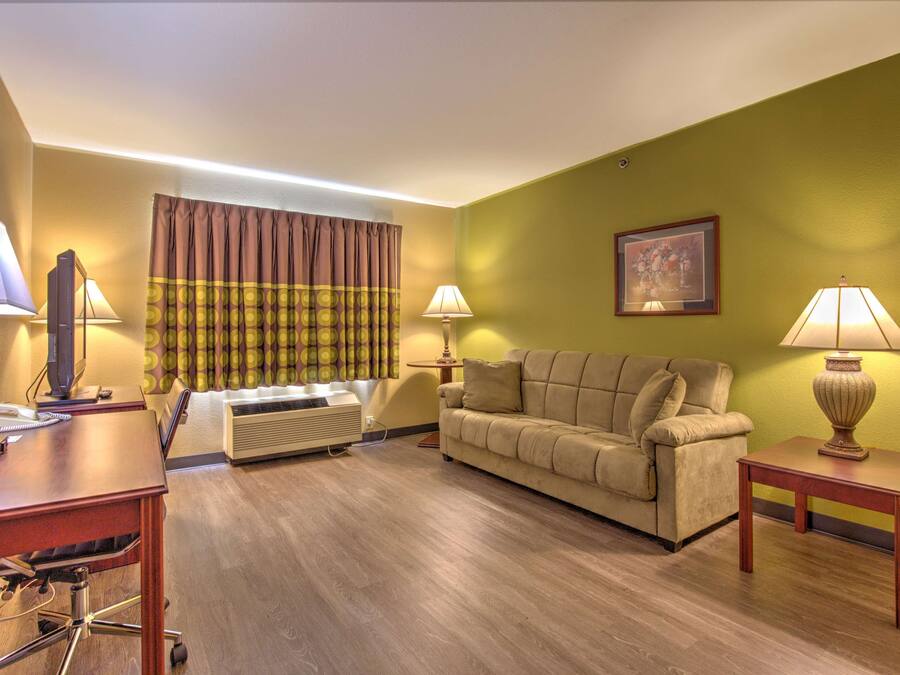A hotel room with a wooden floor, a beige sofa, a desk with a computer and phone, two lamps, and a window with curtains.