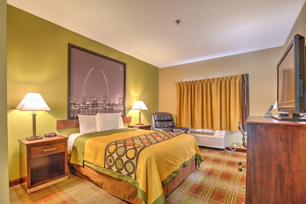 Ein Hotelzimmer mit einem Bett, zwei Nachttischlampen, einem Sessel, einem Schreibtisch mit Computer, einem Fernseher und einem großen Stadtbild an der Wand.
