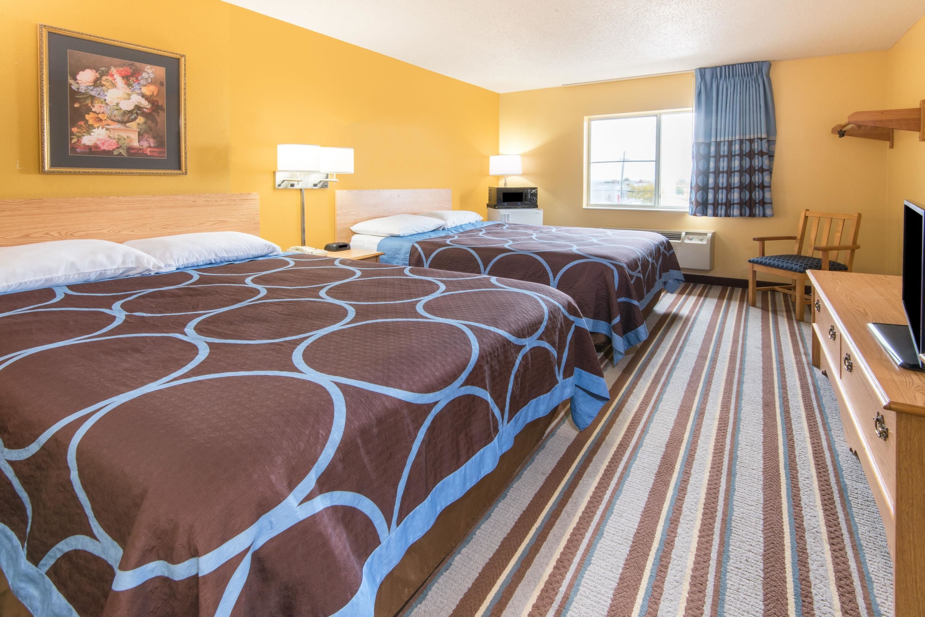 Quarto de hotel com duas camas, uma escrivaninha, uma cadeira, televisão e janela com cortinas.
