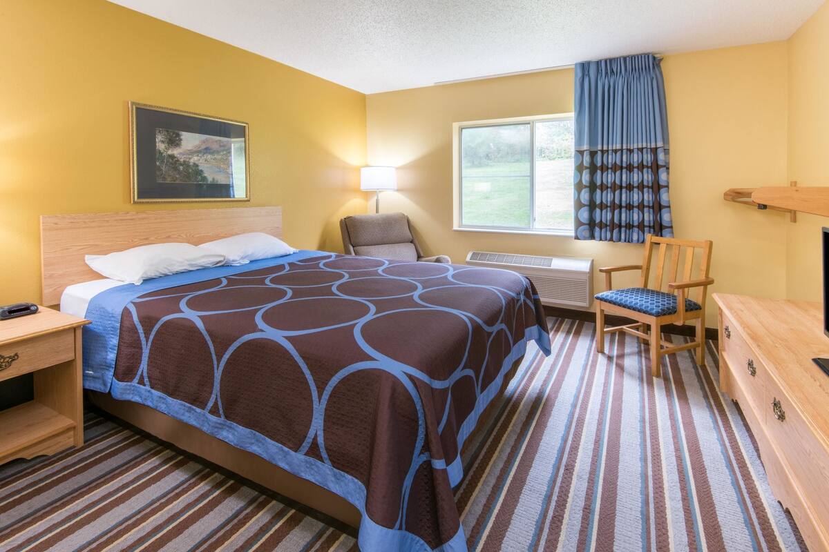 A hotel room with a bed, a chair, a desk, a lamp, a window with curtains, and a picture on the wall.