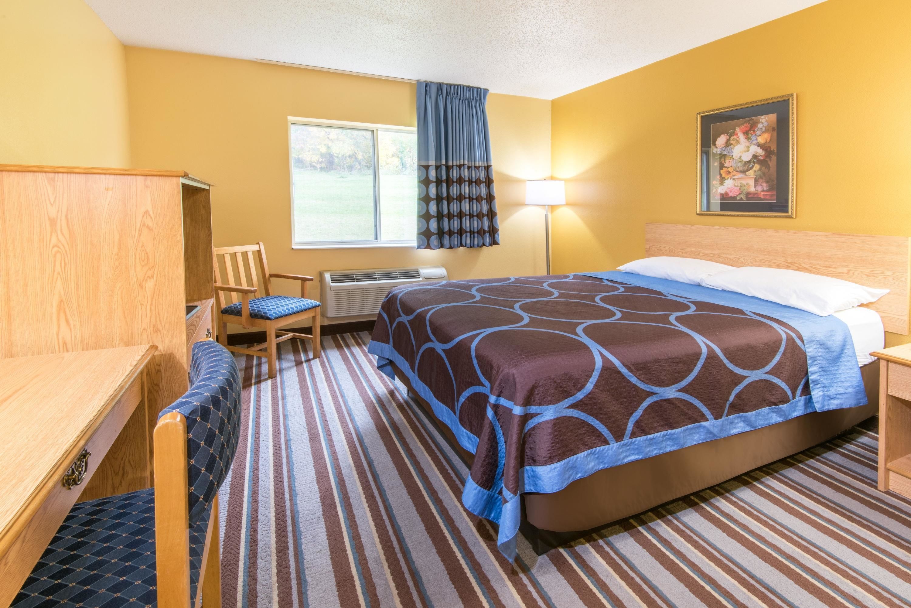 A hotel room with a bed, a desk, a chair, a window with curtains, a lamp, and a painting on the wall.
