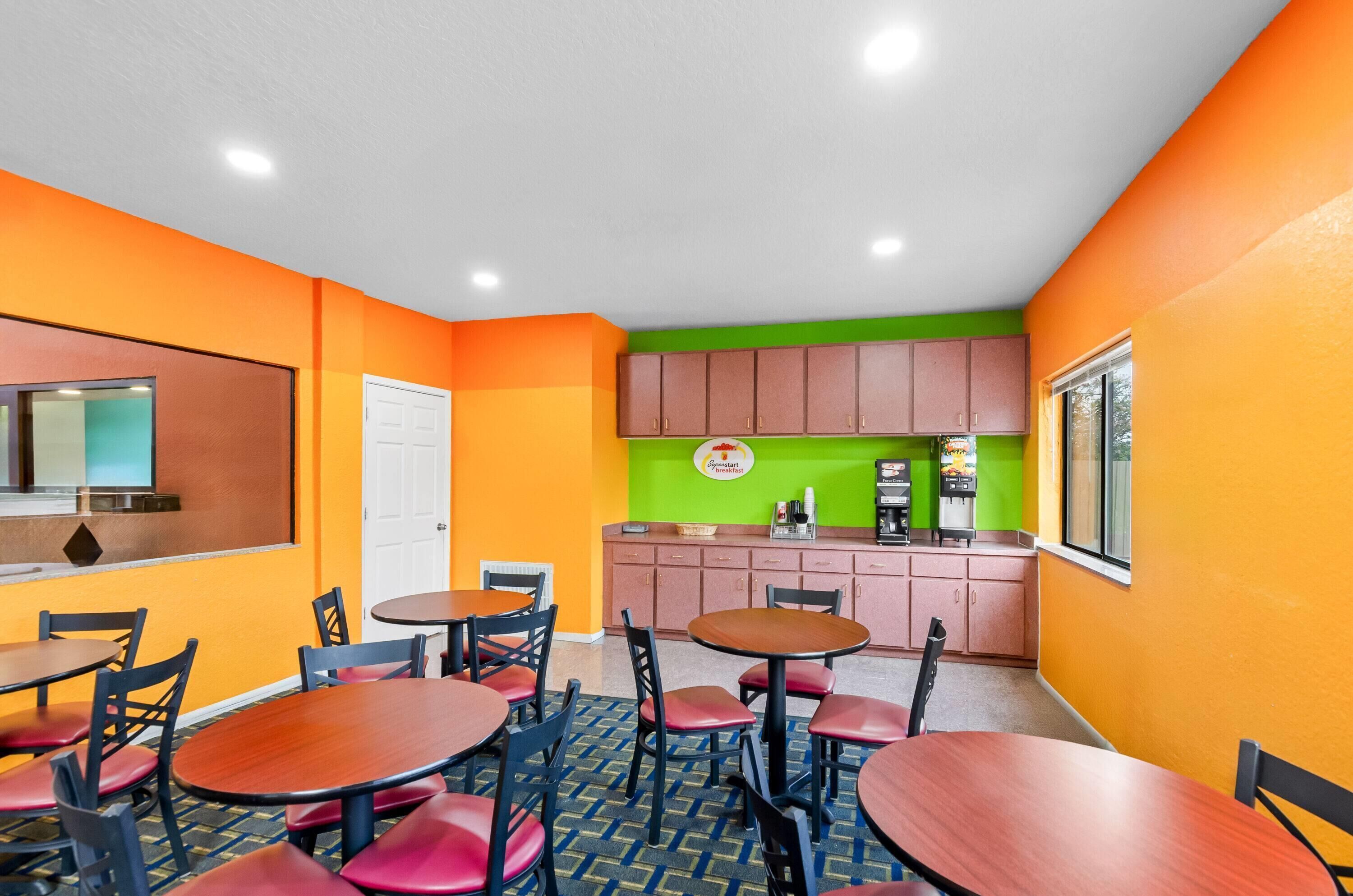 Una habitación de colores vivos con paredes naranjas, una pared de acento verde y una zona de cocina con armarios rosados. Hay varias mesas y sillas dispuestas para comer.