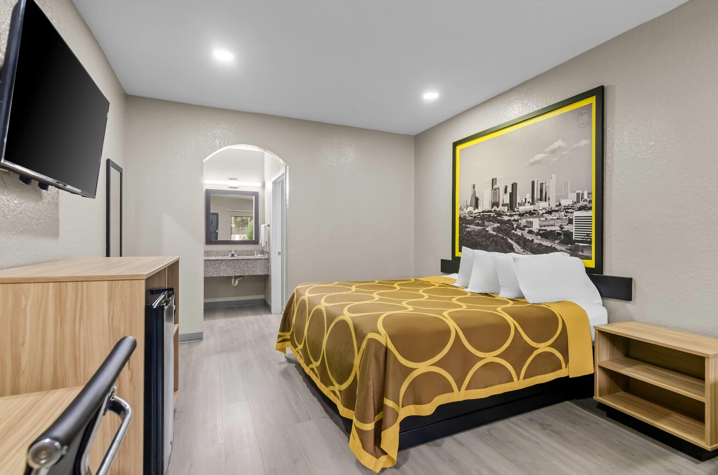 A hotel room with a bed, a desk, a television, and a cityscape painting.