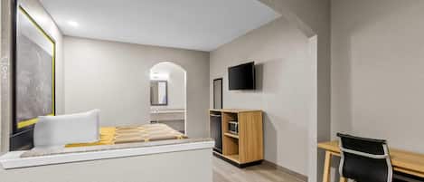 A modern hotel room with a bed, a desk, a chair, a TV, and a small kitchenette.
