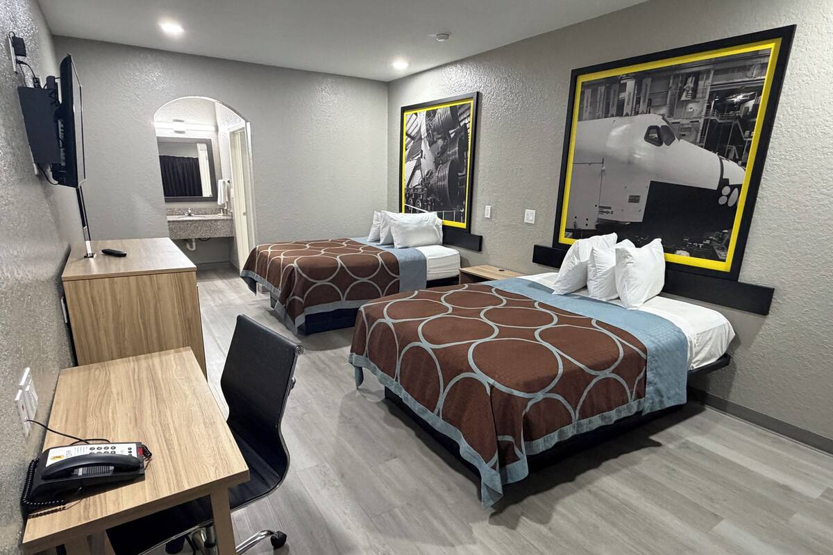 A hotel room with two beds, a desk, a chair, a TV, and a bathroom visible through an arched doorway.
