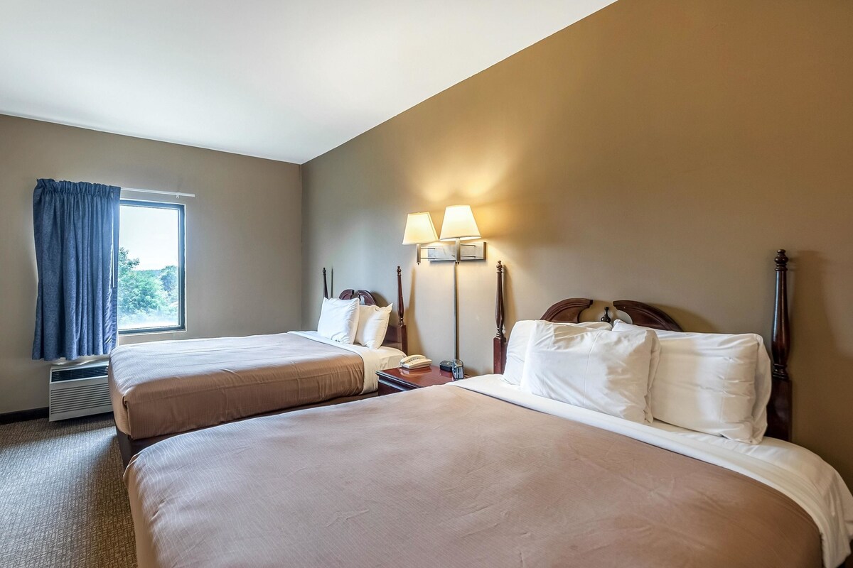 A hotel room with two beds, a window with curtains, a wall-mounted lamp, and a bedside table.
