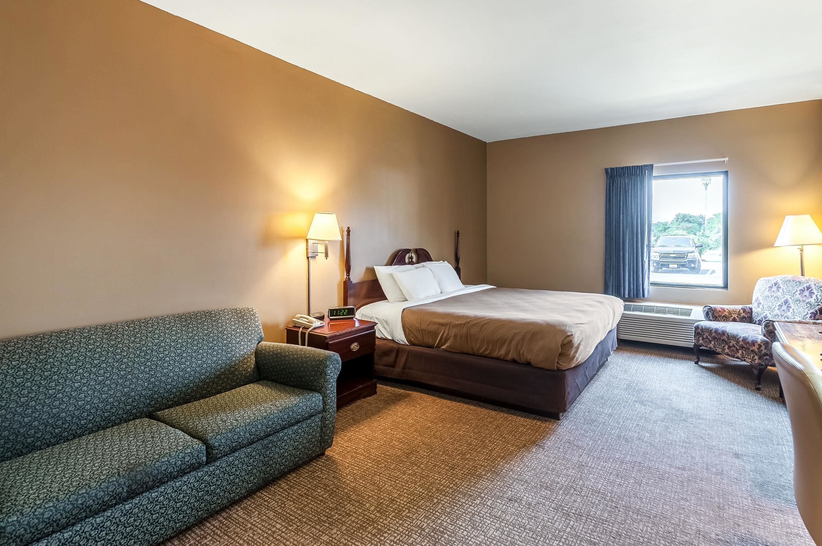 Quarto de hotel com cama, sofá, escrivaninha, luminária e janela com cortinas.
