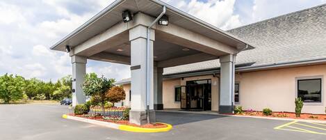 A hotel entrance with a covered walkway, a parking lot, and a building with a grey roof.