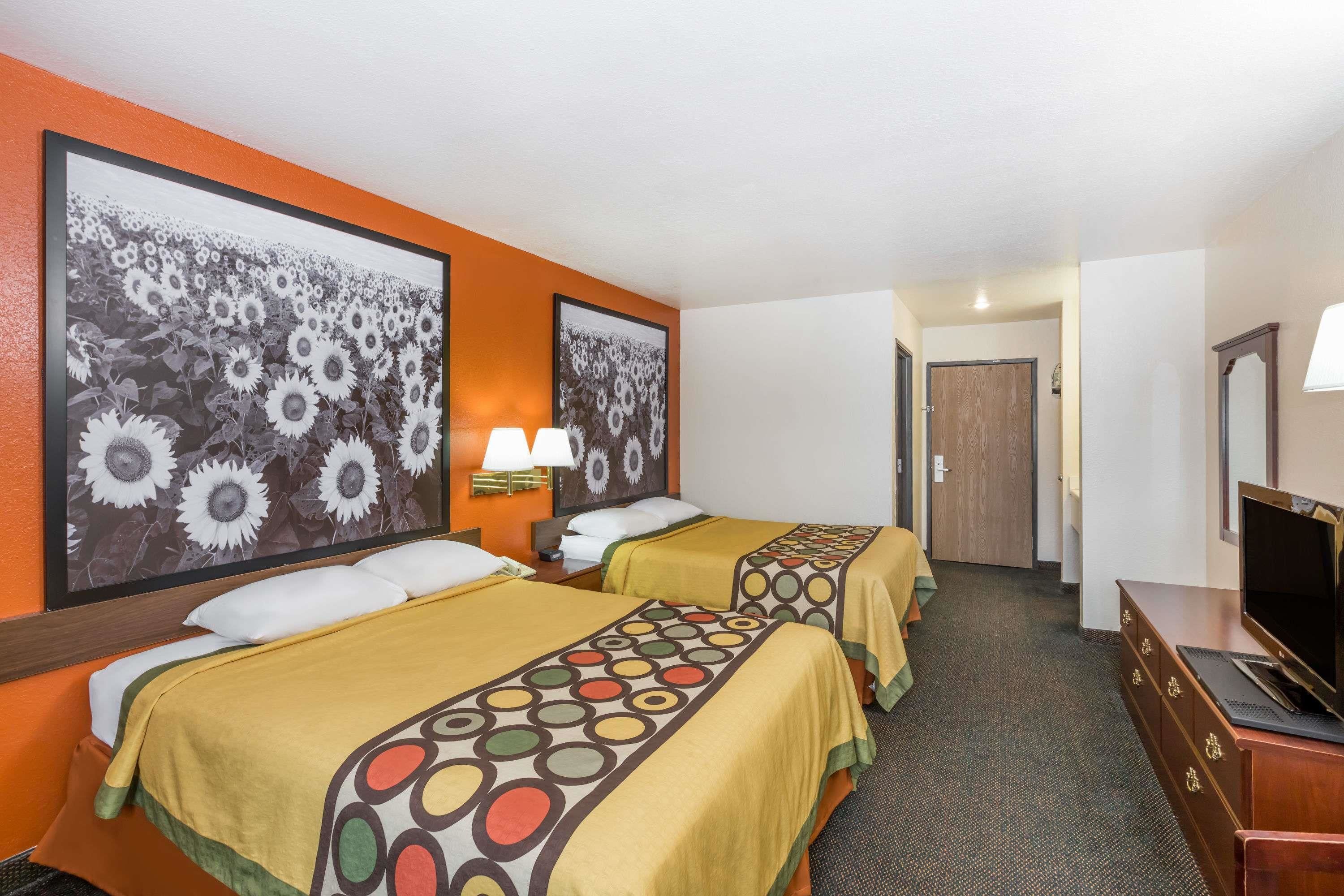 A hotel room with two beds, each with a patterned bedspread, a television on a dresser, and a large sunflower painting on the wall.