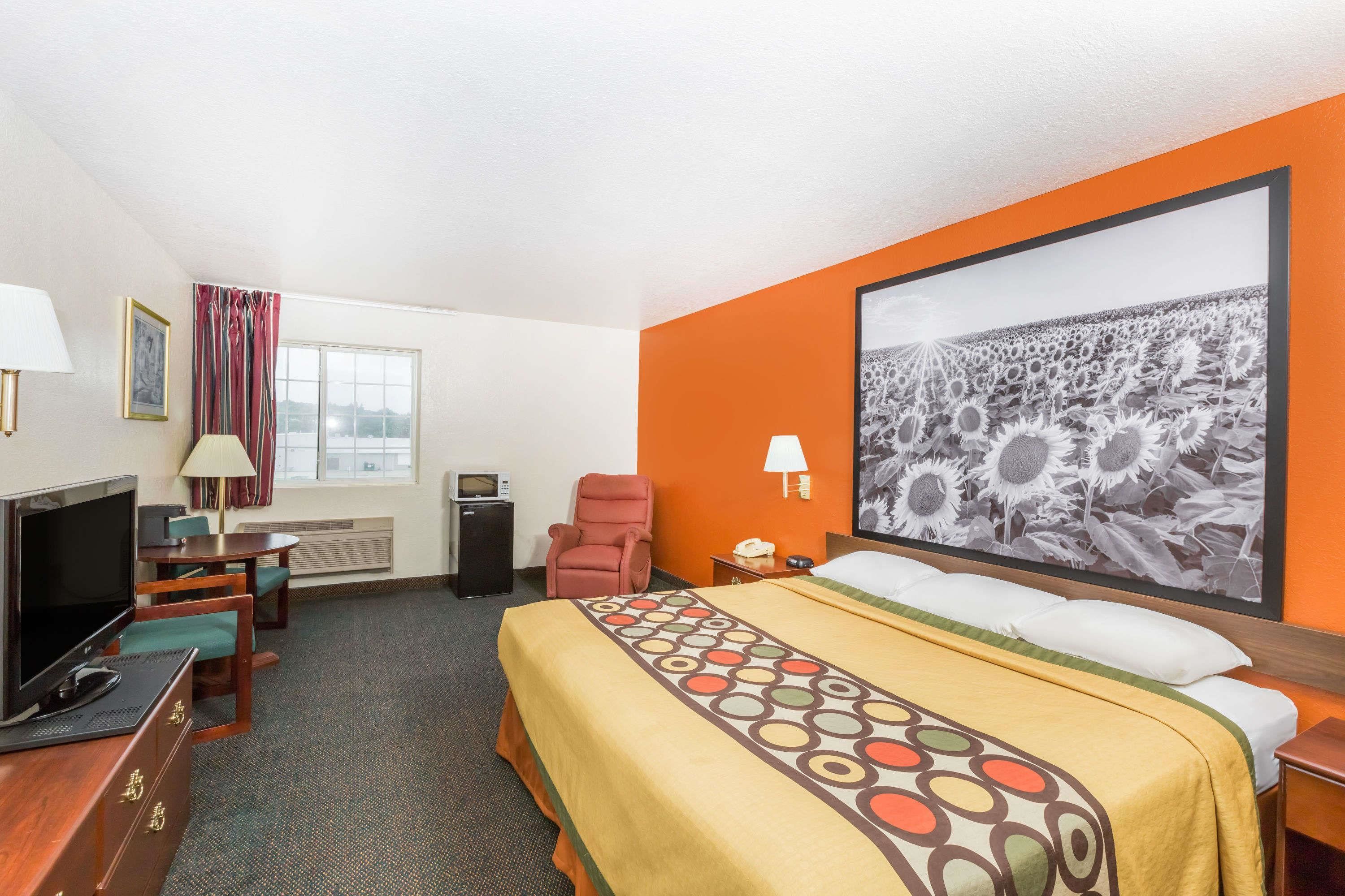 A hotel room with a bed, a TV, a desk, a chair, a desk lamp, a telephone, and a large painting of sunflowers.