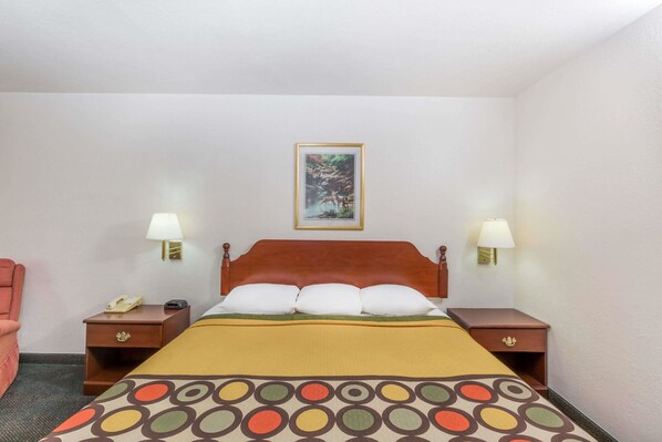 Una habitación de hotel con una cama, dos mesitas de noche, una lámpara, una silla y un cuadro en la pared.