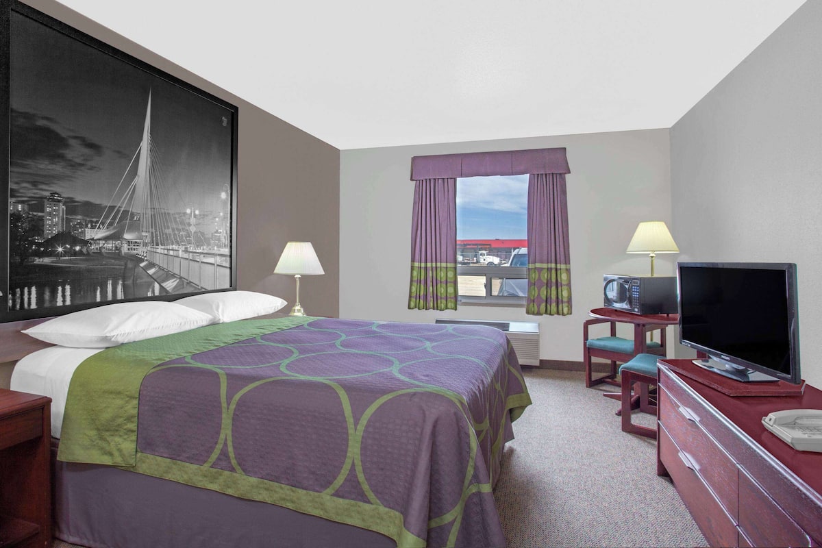 A hotel room with a large bed, a television, a desk with a chair, and a window with curtains.