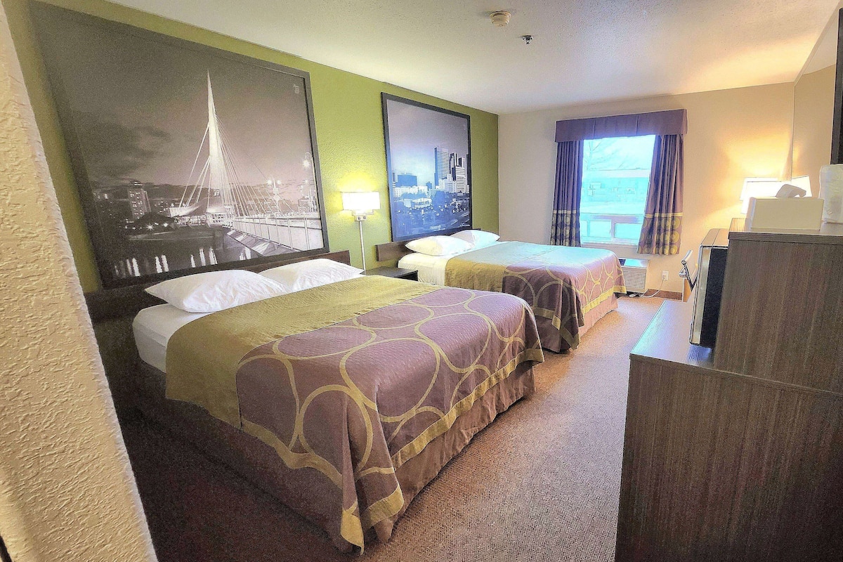 A hotel room with two beds, a desk, a chair, a lamp, and a large window.