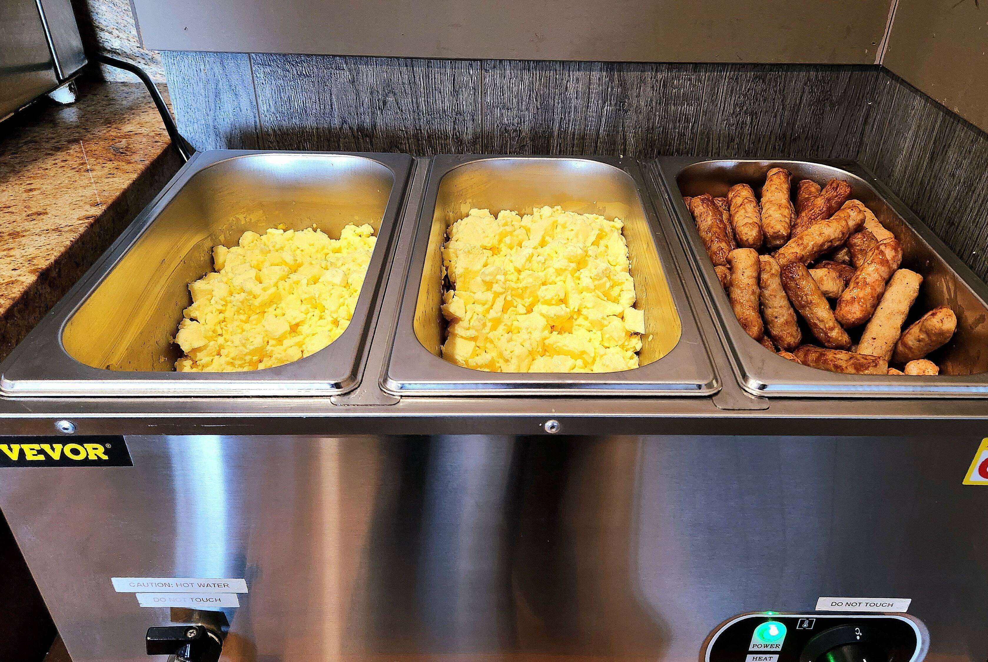 A buffet with three stainless steel trays containing scrambled eggs, hash browns, and sausages.