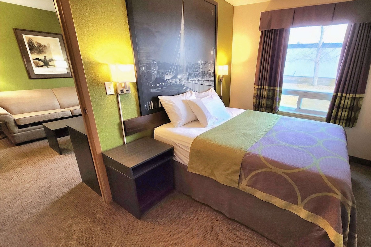 A hotel room with a bed, a nightstand, a desk, a chair, and a view of a cityscape through the window.