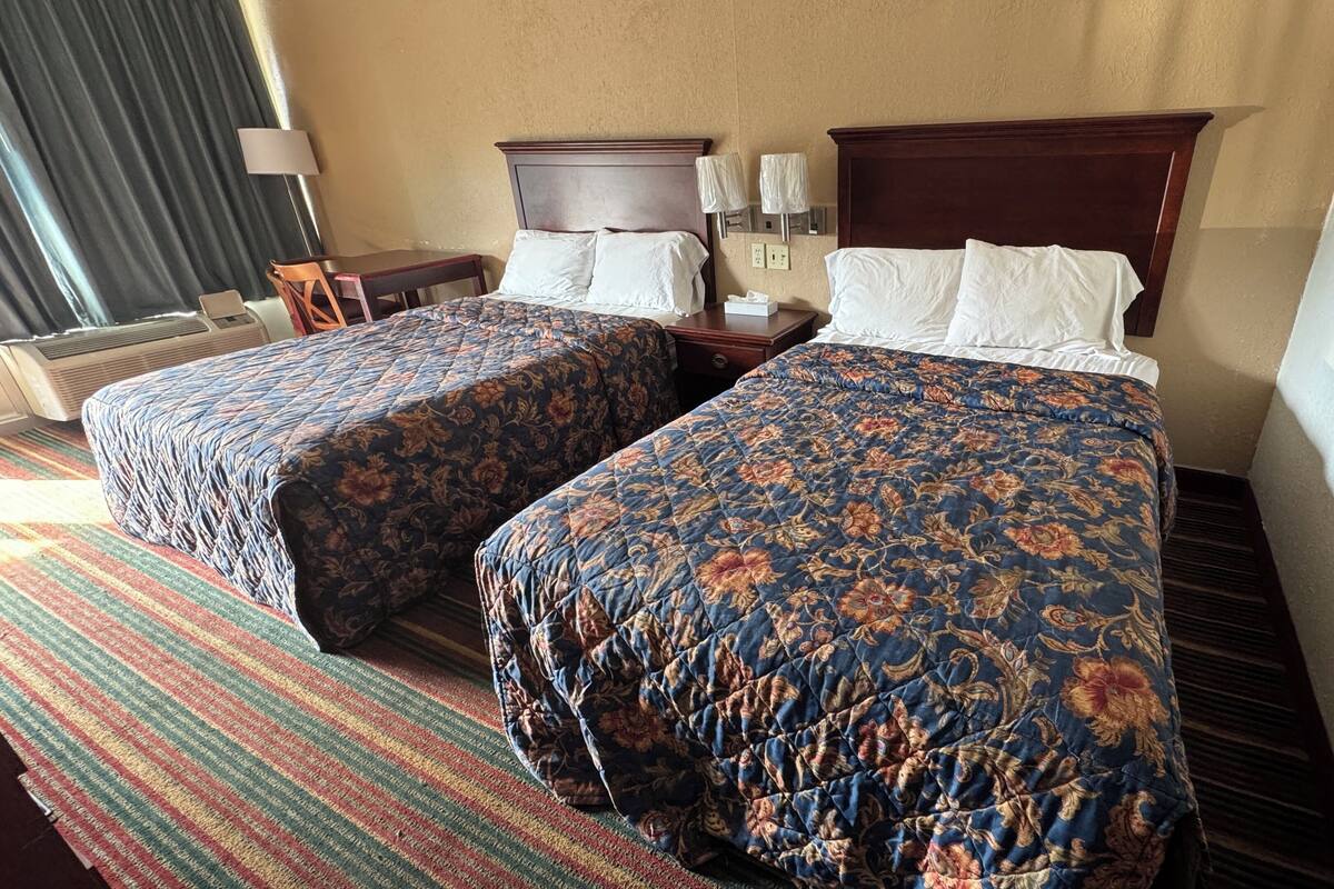 A hotel room with two beds, a desk, a chair, and a lamp.