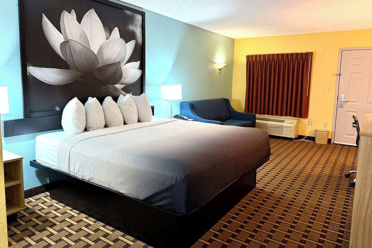 Una habitación de hotel con una cama grande, un sillón azul, una obra de arte en la pared y una ventana con cortinas.