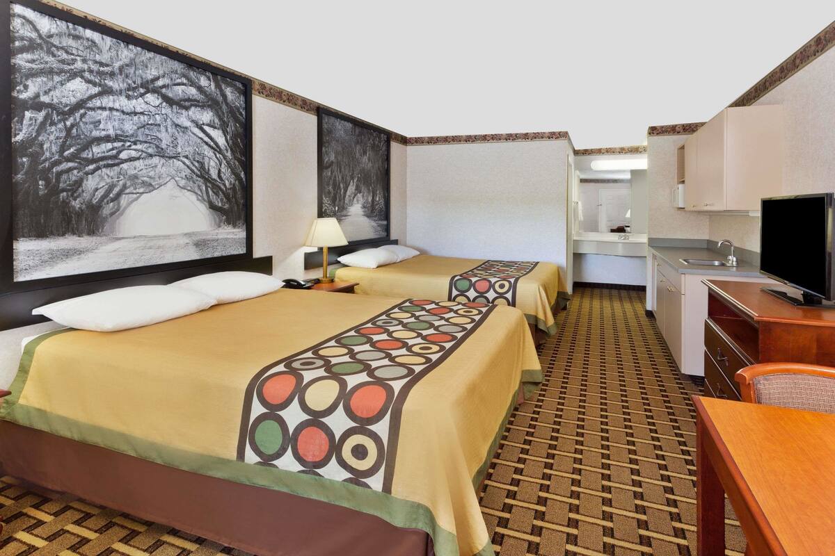 Habitación de hotel con dos camas, un escritorio, un televisor y un ventanal con vista a árboles.