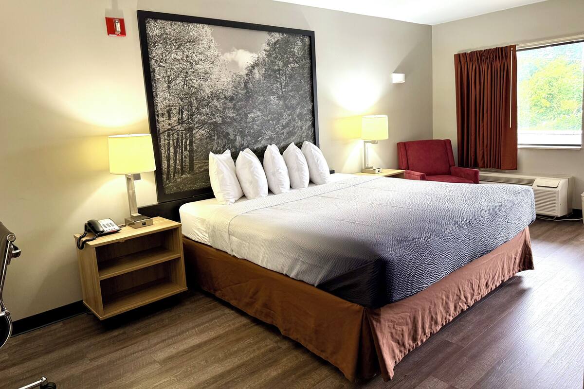 Un cuarto de hotel con una cama grande, dos mesitas de noche con lámparas, una silla roja, un cuadro grande en la pared y una ventana con cortinas.
