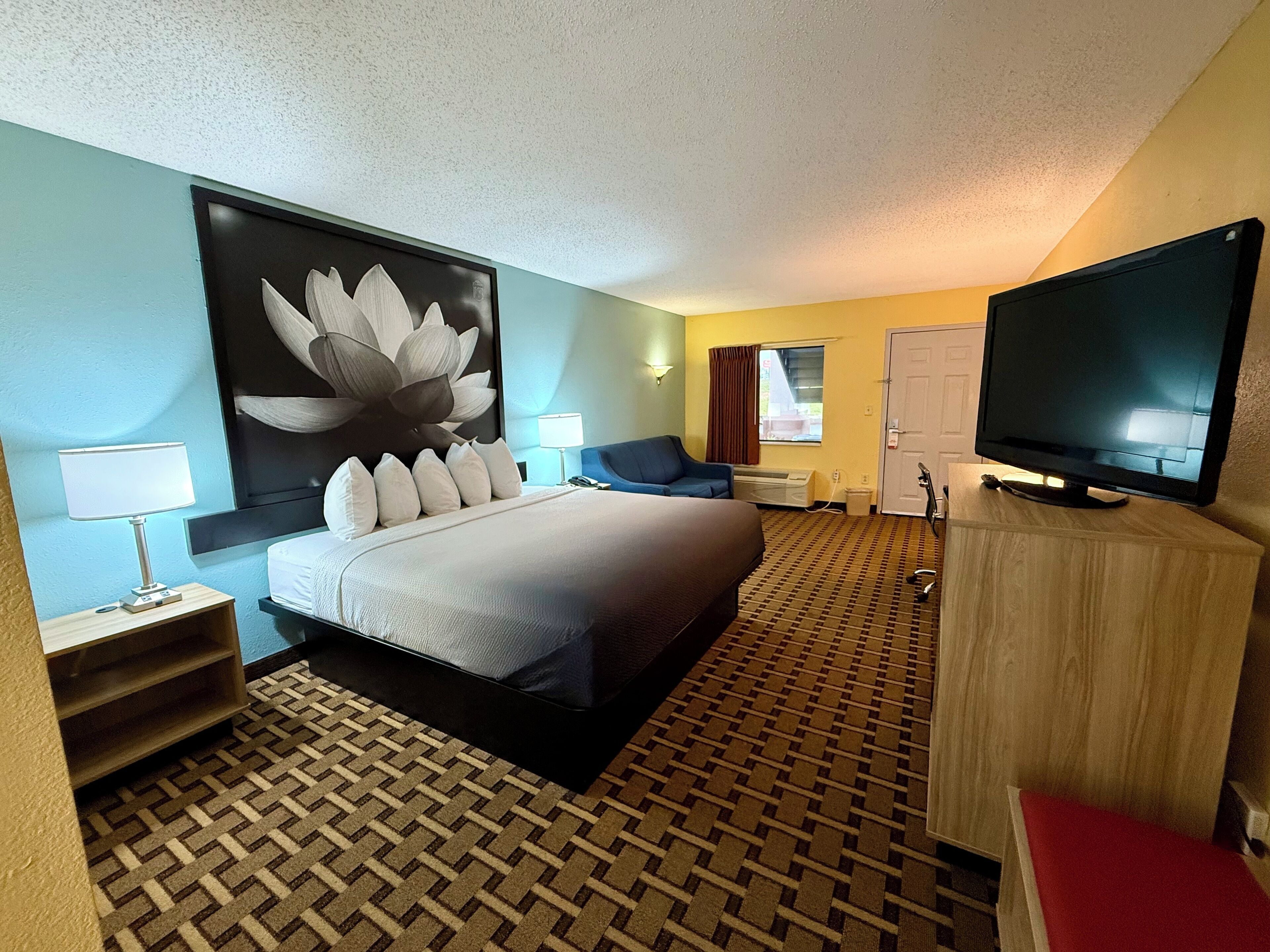 Habitación de hotel con una cama grande, un televisor de pantalla plana, un escritorio con lámpara, una silla y una obra de arte colgada en la pared.