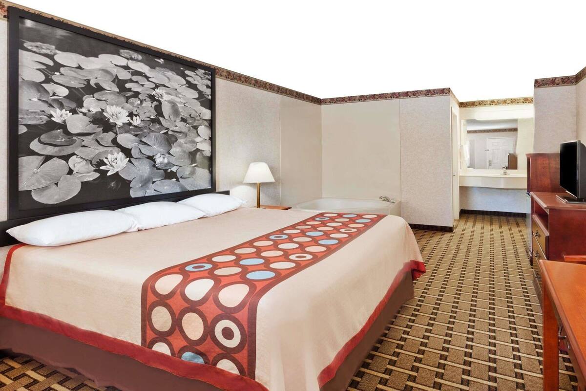 Habitación de hotel con cama, un cuadro grande enmarco con nenúfares, una lámpara, televisión y un baño visible a través de una puerta abierta.