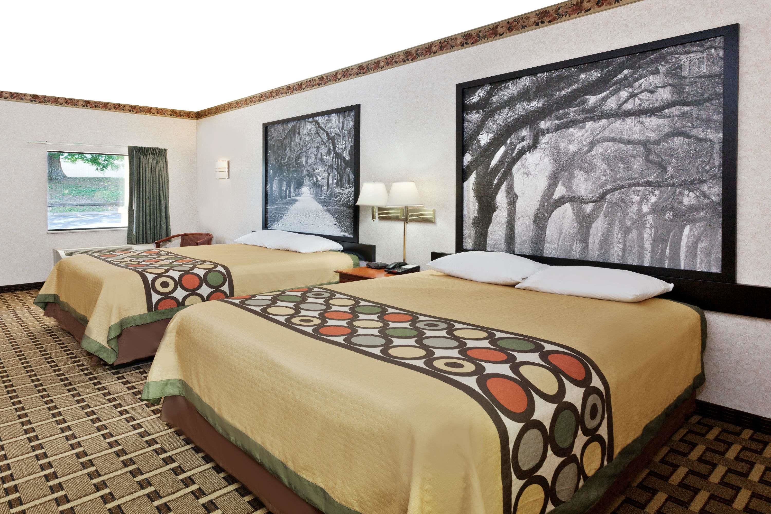 A hotel room with two beds, each with a patterned bedspread, a large window with curtains, and two framed pictures on the wall.
