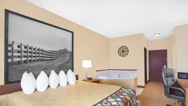 A hotel room with a large bed, a desk, a chair, a desk lamp, a wall-mounted TV, a large framed picture, and a bathroom with a bathtub and a toilet.