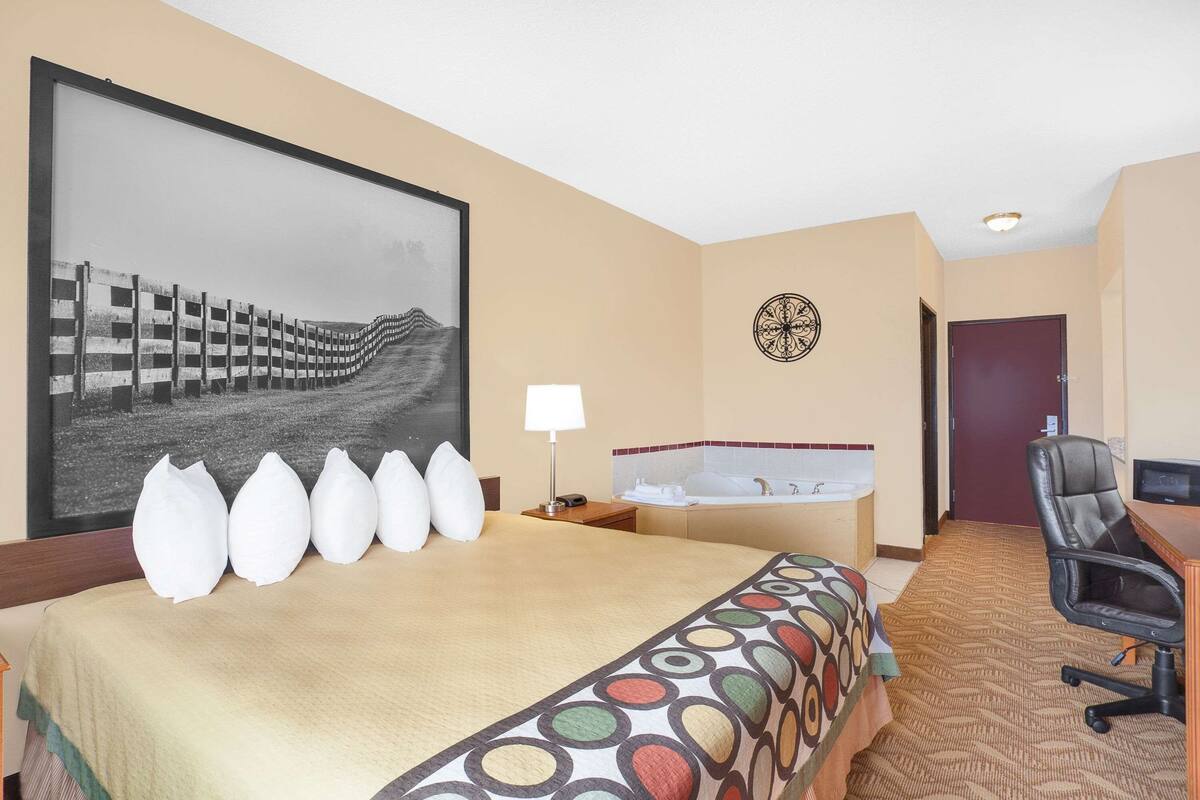 Hotelkamer met een groot bed, een bureau, een stoel, een bureaulamp, een aan de muur bevestigde televisie, een groot gepresenteerd schilderij, en een badkamer met een bad en een toilet.