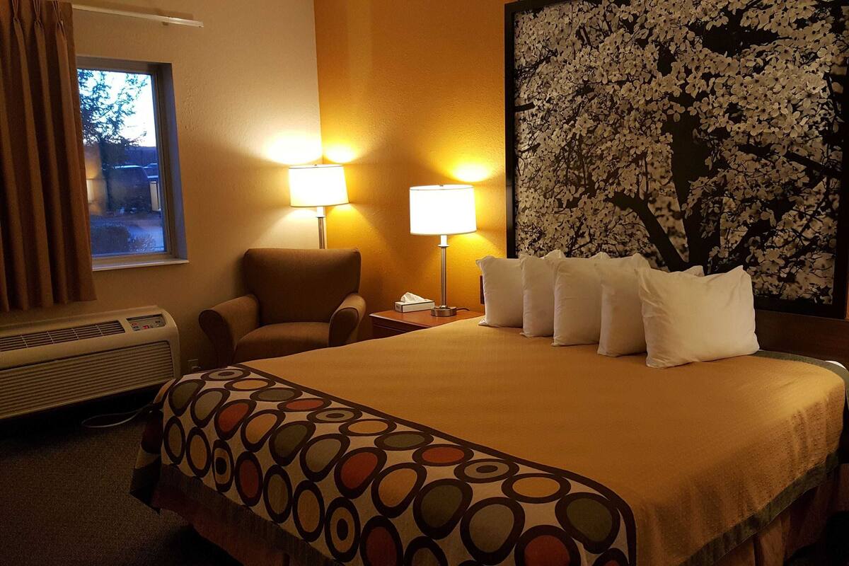 Een hotelkamer met een bed, twee bedlampen, een stoel, een groot bloemenkunstwerk aan de muur en een raam met gordijnen.