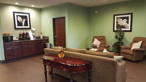 A living room with a sofa, two armchairs, a coffee table, and a wooden side table. There is a kitchen area with a microwave, coffee maker, and a basket of oranges.