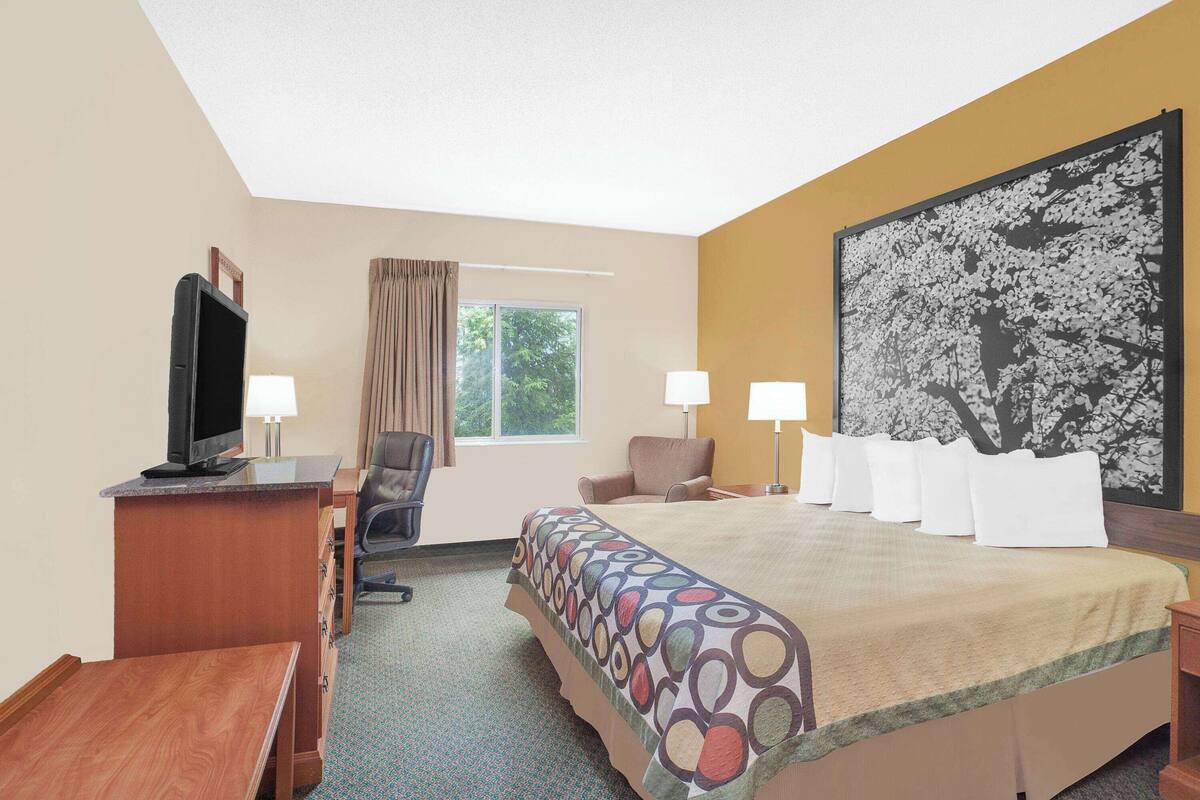 Een hotelkamer met een bed, een bureau met een televisie, een stoel, een raam met gordijnen, en een groot bloemen schilderij aan de muur.
