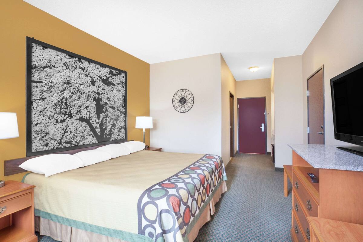 Een hotelkamer met een bed, een televisie, een dressoir, een klok en een deur naar een andere kamer.
