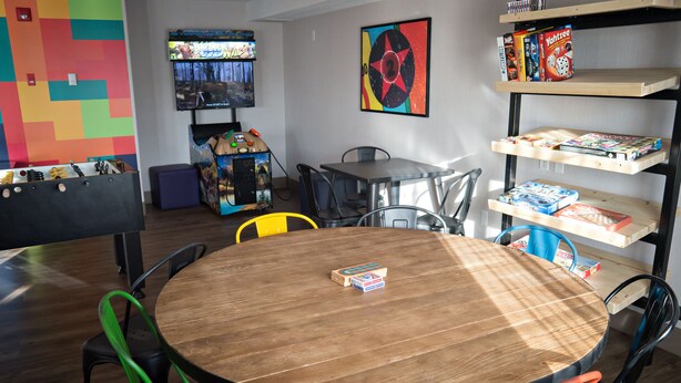 A room with a wooden round table, green and orange chairs, a TV, a colorful wall, and shelves with board games. - Canalta Hotel St Paul (St. Paul)