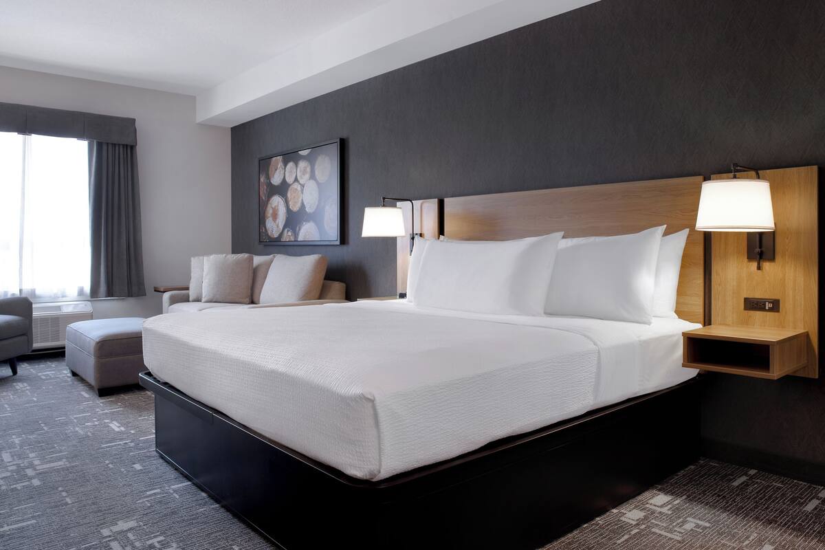 A modern hotel room with a large bed, a bedside table with a lamp, a chair, and a small table.