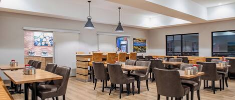 A modern dining area with wooden tables and leather chairs, a brick wall, and a large window.