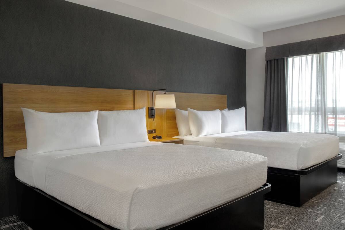 A hotel room with two beds, a wooden headboard, a nightstand with a lamp, and a large window with curtains.