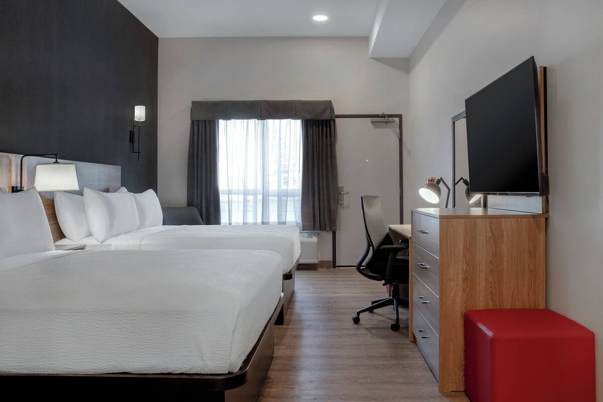 A hotel room with two beds, a desk with a chair, a TV, and a red ottoman.