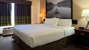 A hotel room with a large bed, a nightstand, a lamp, a window with curtains, and a painting of a landscape.