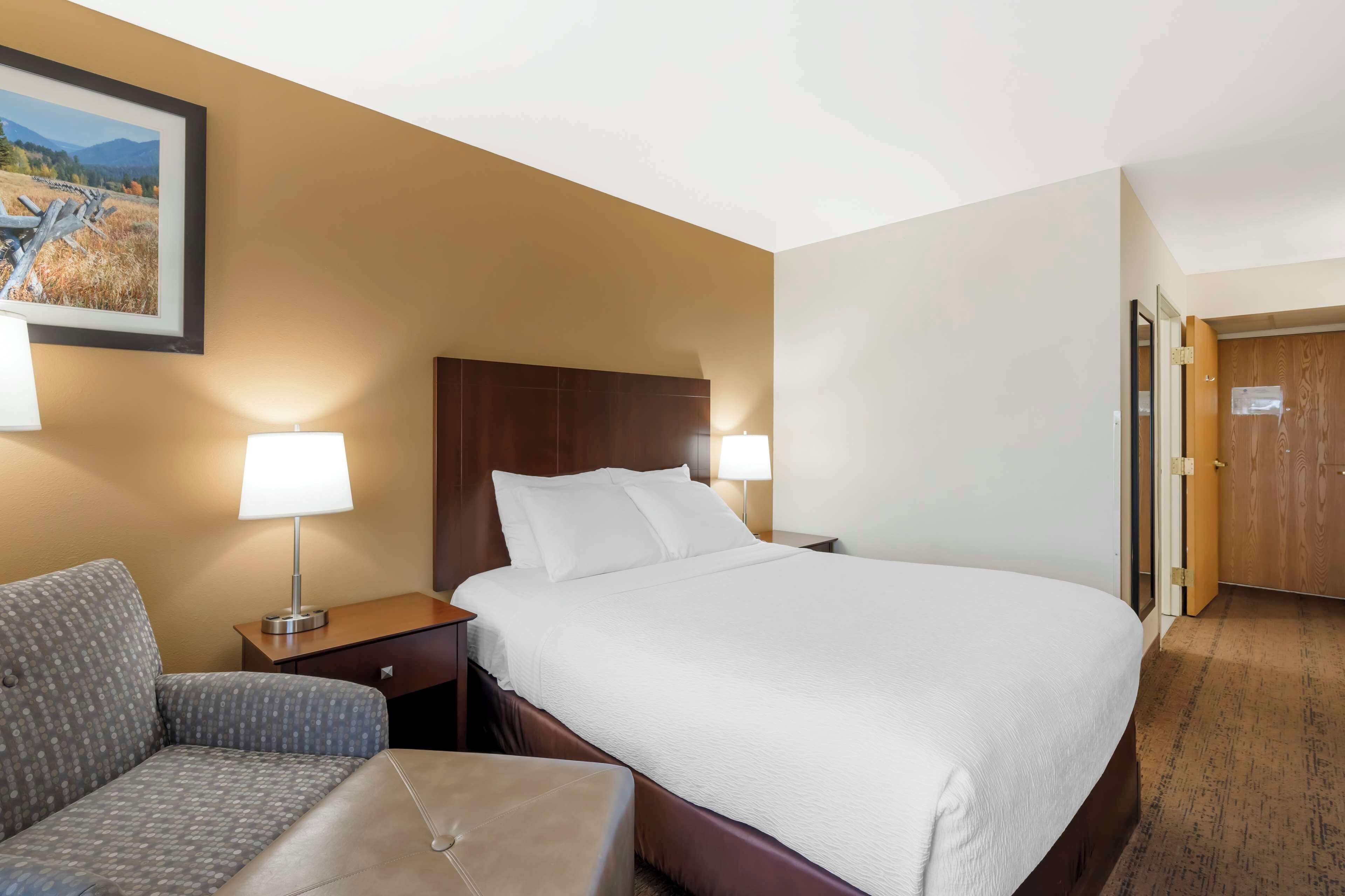 Una habitación de hotel con una cama grande, una silla, una mesita de noche con lámpara y una puerta de madera.