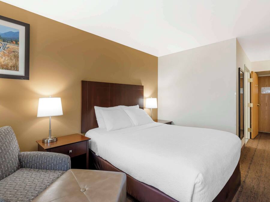 Una habitación de hotel con una cama grande, una silla, una mesita de noche con lámpara y una puerta de madera.