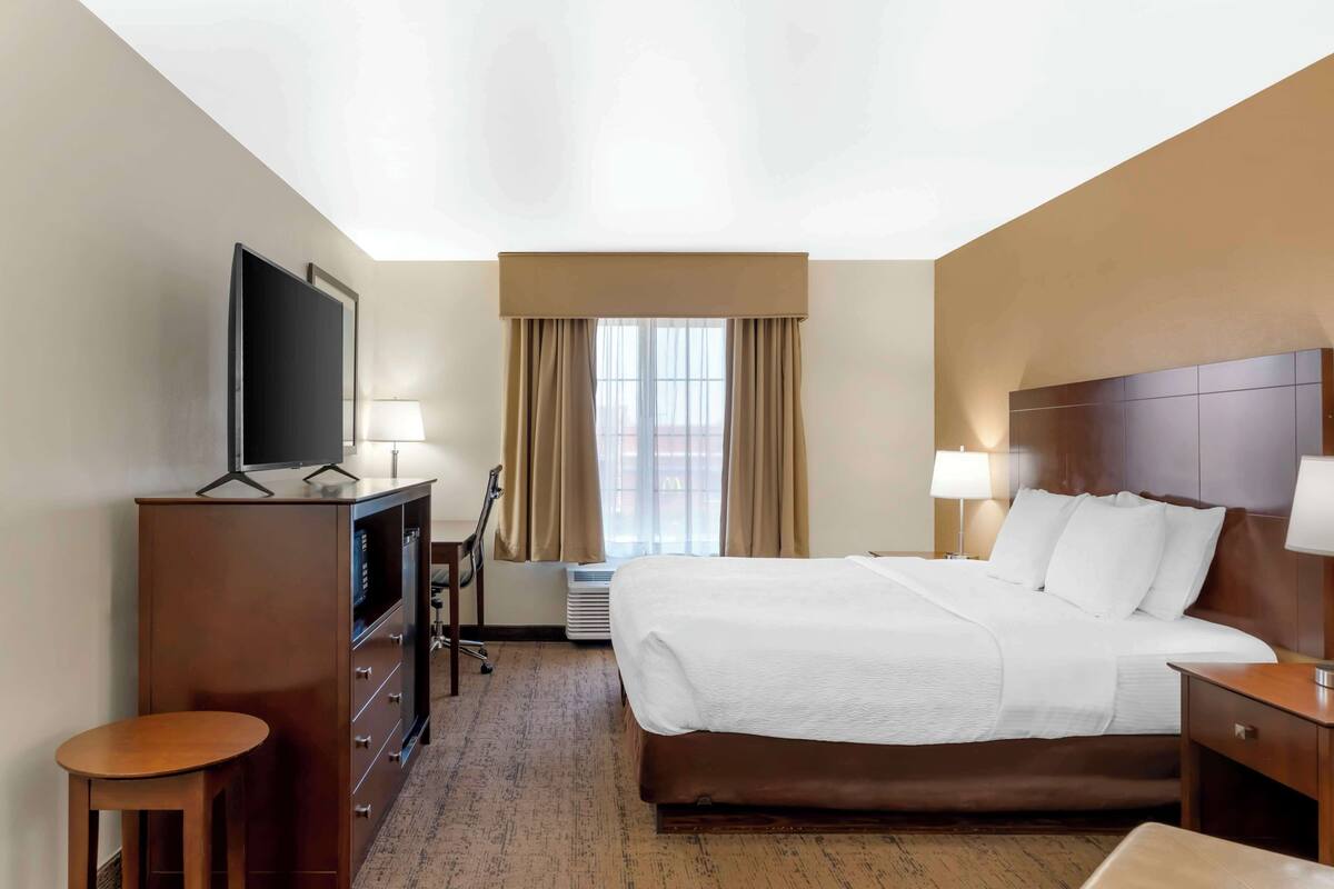 A hotel room with a large bed, a TV on a dresser, a desk, and a window with curtains.