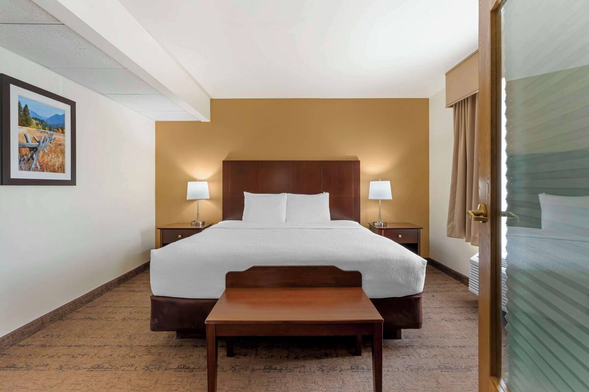 A hotel room with a large bed, two bedside lamps, a wooden nightstand, a small table, and a framed picture on the wall.