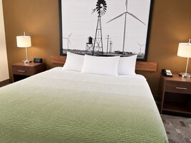 Habitación de hotel con una cama grande, dos mesitas de noche, un reloj, un teléfono y un cuadro colgado en la pared con aerogeneradores.
