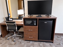 Habitación de hotel con escritorio de madera, una silla de oficina negra, televisor de pantalla plana, microondas, mini refrigerador y cafetera.