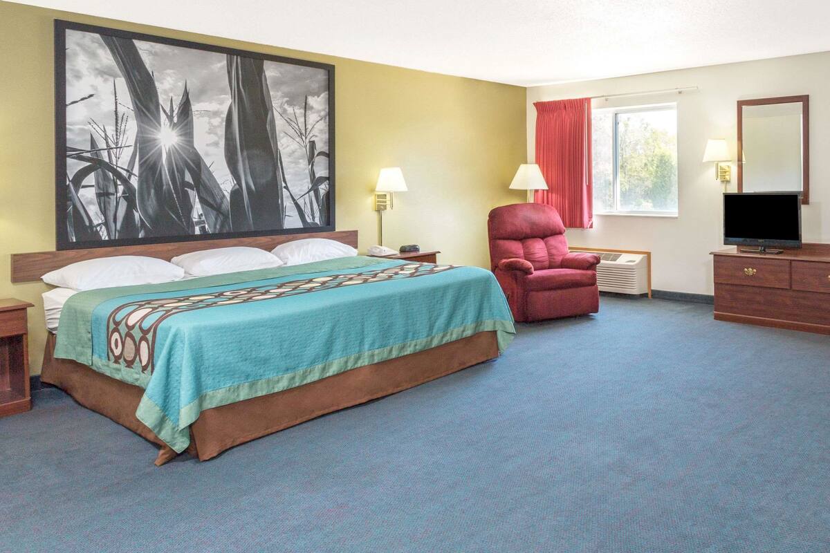 A hotel room with a bed, a red armchair, a TV, and a large painting on the wall.