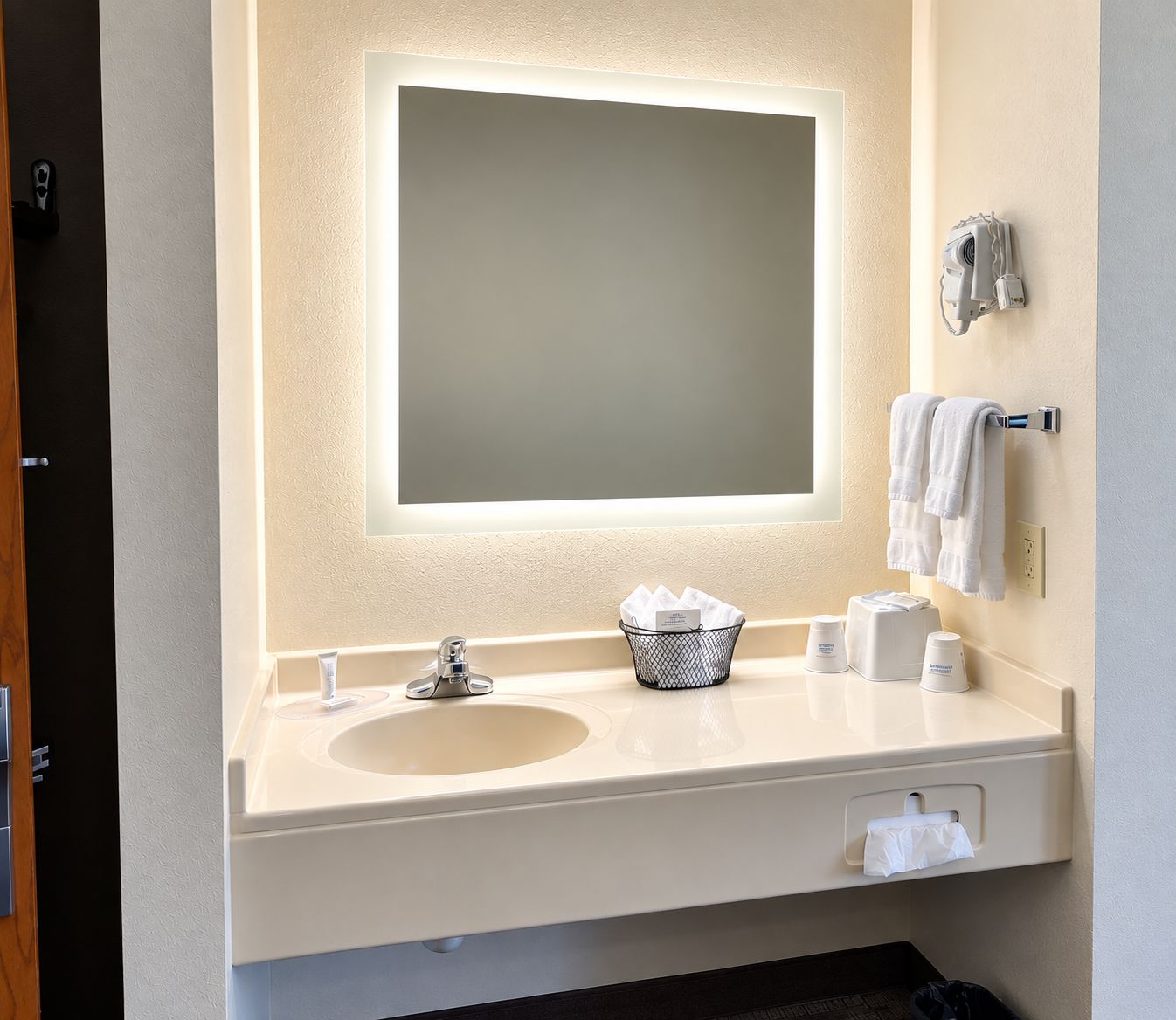 Une salle de bain d’hôtel avec un grand miroir, un lavabo, un porte-serviettes et un distributeur de mouchoirs.