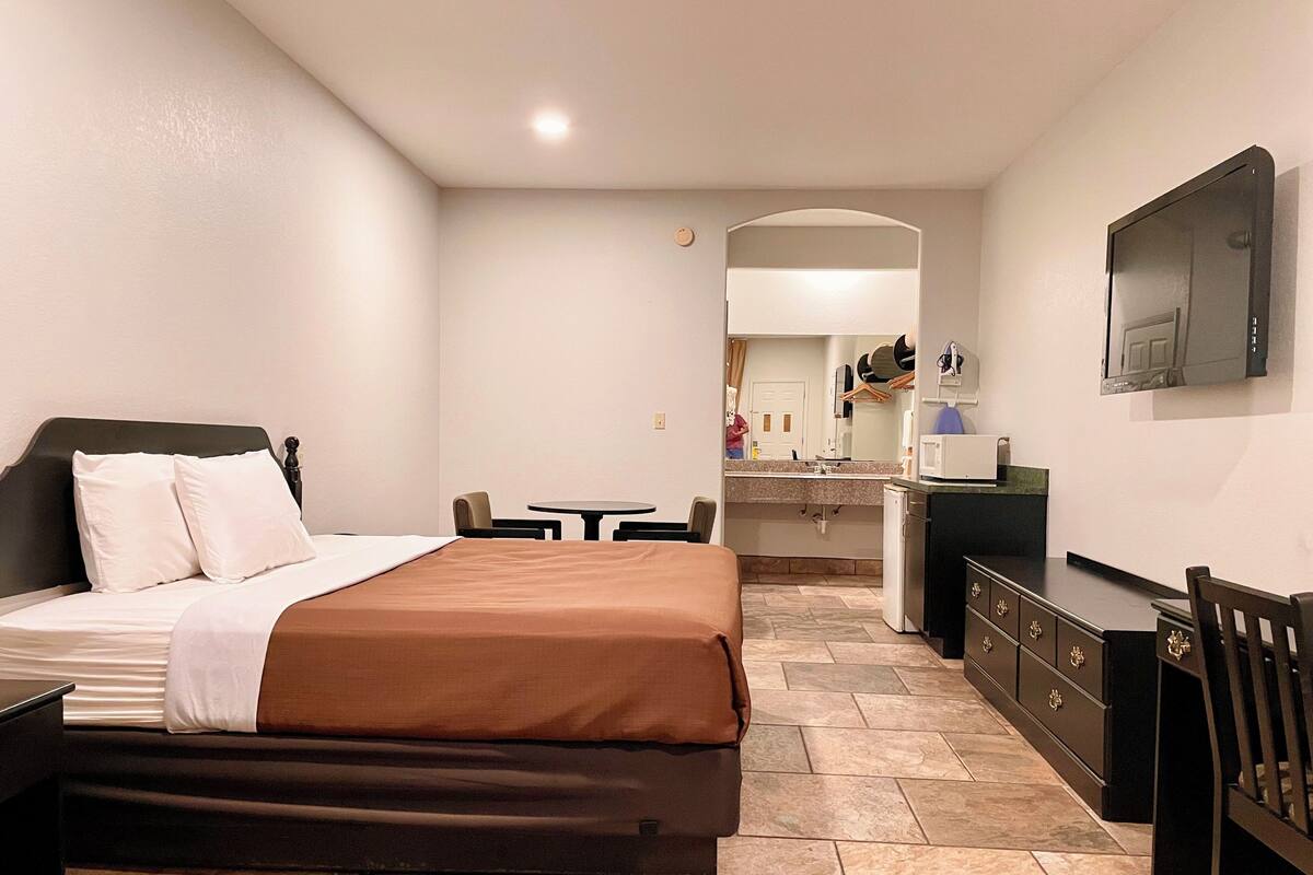A hotel room with a bed, a desk, a chair, a small table, a mirror, and a television.